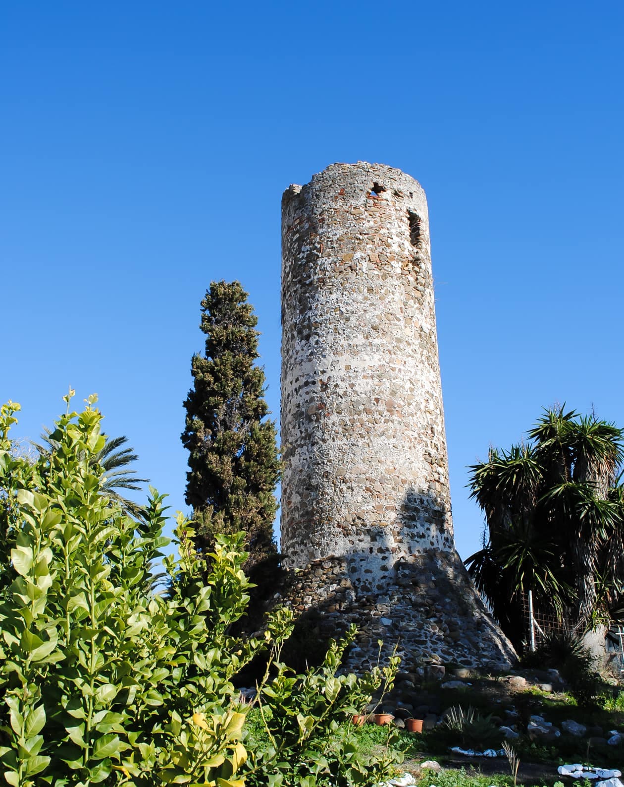 Torre almenara de Casasola o Baños, de mampostería de cantos rodados y base ataludada, entre cipreses y palmeras