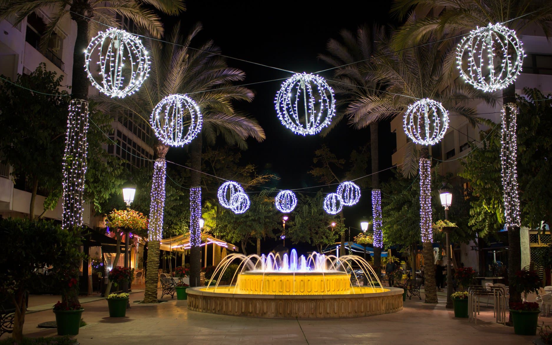 Bolas de luz navideñas colgadas entre palmeras sobre una fuente iluminada en el centro de Estepona