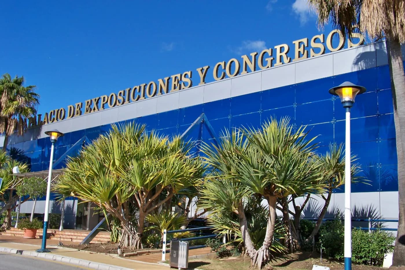 Fassade des Palacio de Exposiciones y Congresos in Estepona mit goldenen Buchstaben über blauem Glas und Palmen davor